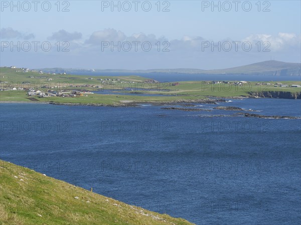 Extensive coastal landscape with houses and hills bordering the blue sea, green meadows on a deep
