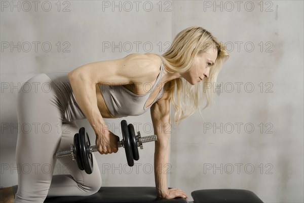 Muscular middle-aged woman lifting a dumbbell while leaning on a sports bench, view in profile