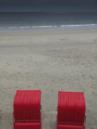 Two red beach chairs stand on an empty beach in front of a calm sea under a cloudy sky, beach