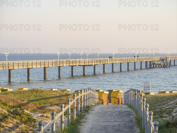 A long jetty leads into the sea, next to it beach chairs and a yellow sky at sunset, beach chairs