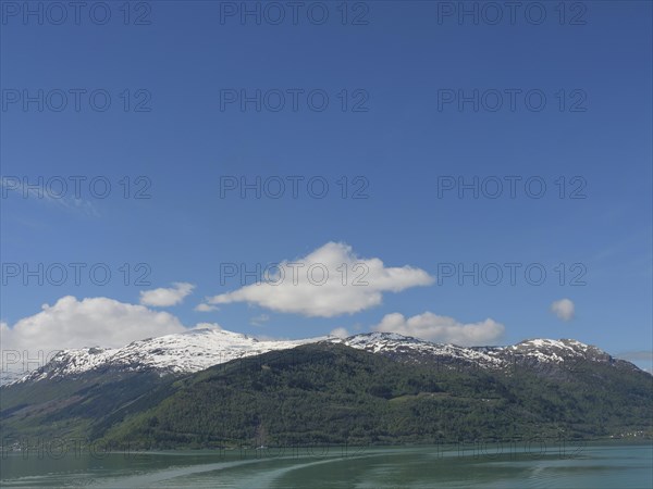A mountain with snow peaks under a blue sky with clouds, in front of a calm lake, greenish