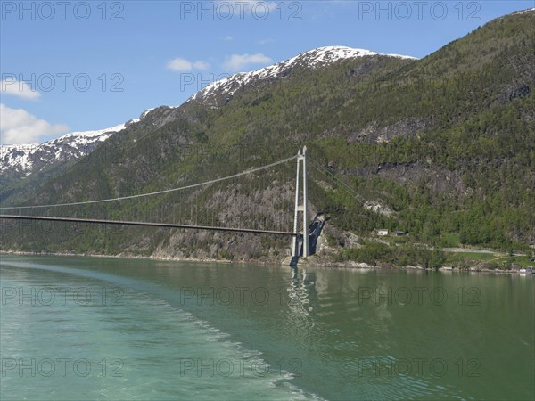 Vast landscape with a bridge stretching over a calm lake and snow-capped mountains under a blue