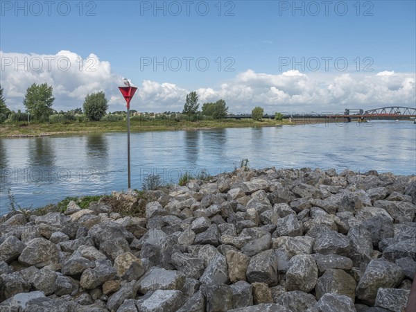 A calm river under a cloudy sky, surrounded by green nature and a bridge in the background, stone