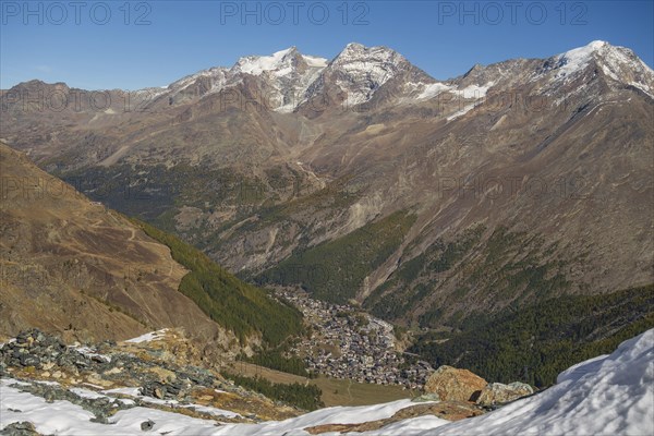 View down the valley to a village surrounded by snow covered mountains under a bright blue sky,