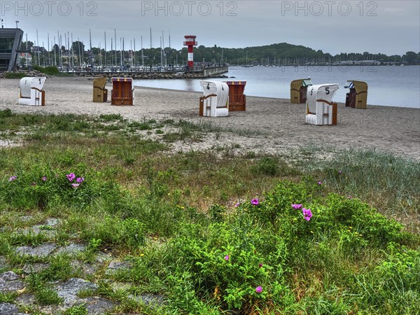 Scattered beach chairs on a sandy beach with a harbour and sailing boats in the background. The sky