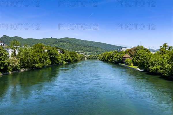 A calm river flows through a landscape of green trees and hills under a blue sky, Olten, Solothurn,