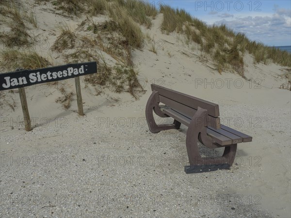 A bench by the sand dunes, next to it a sign with the inscription 'Jan Sietsepad', on it a partly