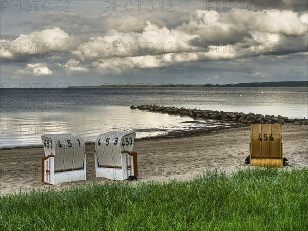 Two white and one yellow beach chair on a sandy beach, rocks in the water and cloudy sky, beach