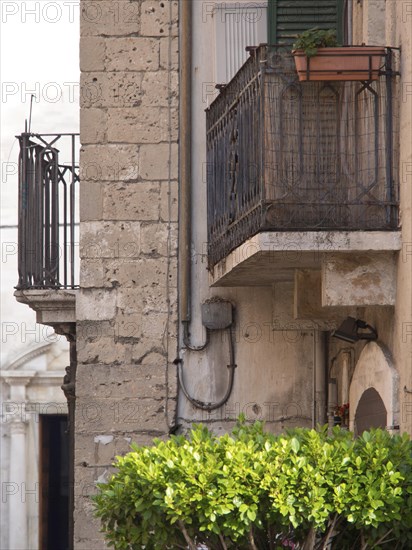 An old balcony on a stone facade with a flower pot and a wild green bush in the foreground, small