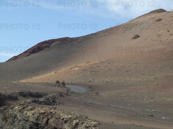 A rocky volcano with a narrow road, surrounded by lava and ash under a slightly cloudy sky, barren