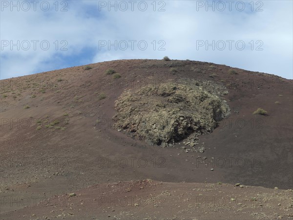 A volcanic slope with lava rocks, light vegetation and partly cloudy sky, barren landscape with