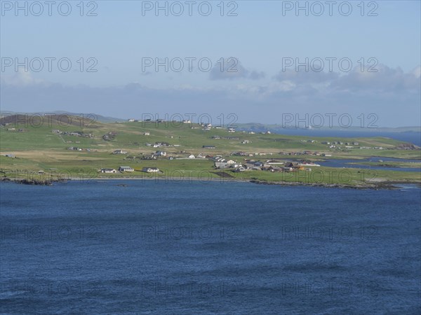 Small settlement with white houses stretches along the grassy coastline in front of the blue sea,