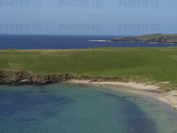 Green grassy landscape along a calm bay with clear blue water under a wide sky, golden beach with