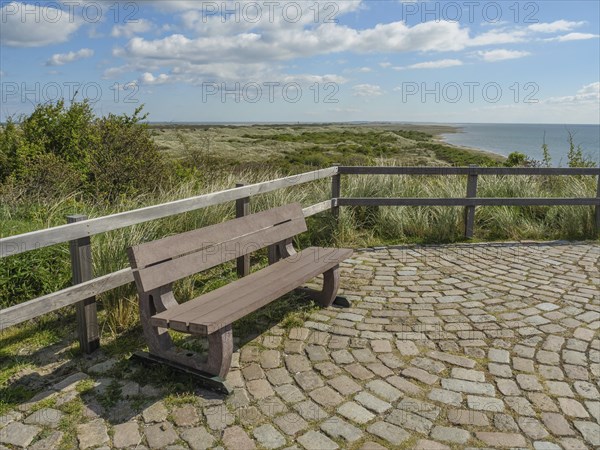 Bench on a paved floor with a view of the sea and the dunes, under a blue sky with clouds, dune and