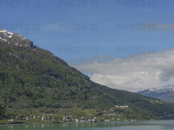 Sunny landscape with snow-covered mountains and houses on the lakeshore under a blue sky, greenish