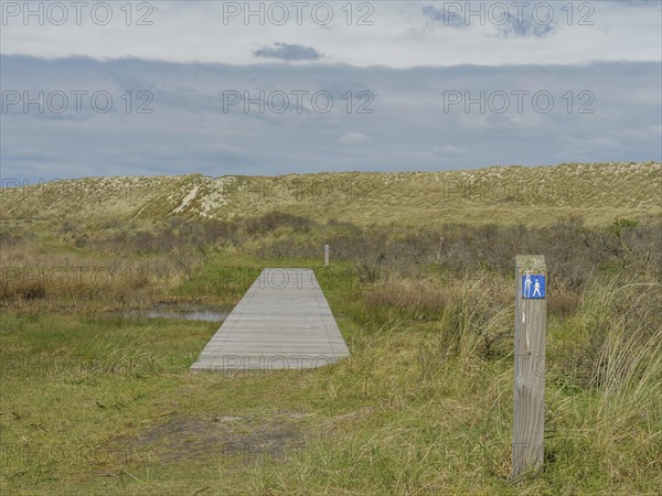 A wooden walkway leads into a dune landscape, next to it a post with a blue sign under a cloudy