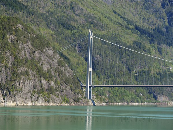 Imposing bridge over bright green water, framed by steep, forested mountains under a blue sky,
