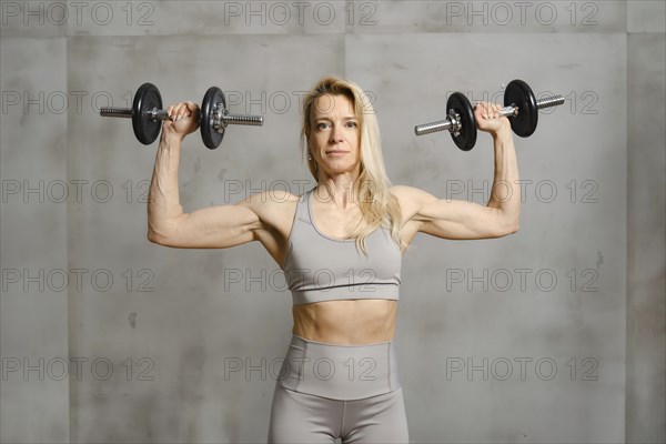 A middle-aged woman stands near the grey wall of the gym and pushing dumbbells up