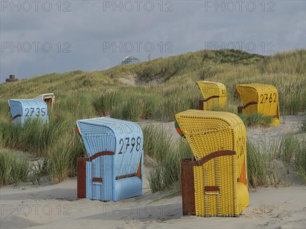 Different coloured beach chairs on the sandy beach, surrounded by dunes and under a cloudy sky,