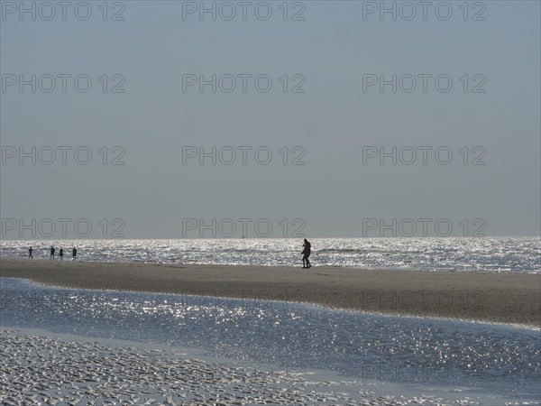 Single person walking along a wide beach, other people visible in the distance, glittering sea