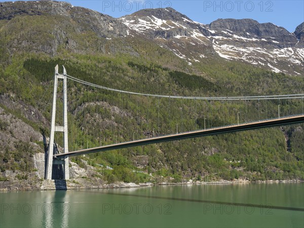 Large bridge over calm water, framed by forested mountains and rocks under a bright blue sky,
