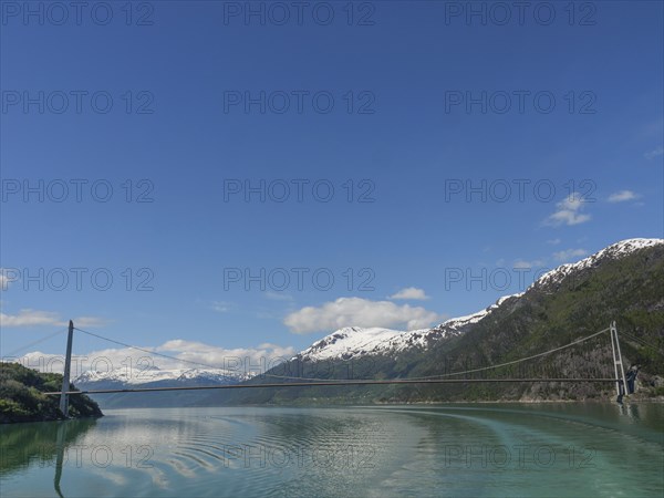 A bridge spans the turquoise water, surrounded by snow-capped mountains under a blue sky, bridge in