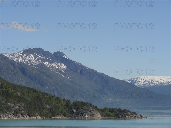 View of steep, forested mountains, a lake and snow-capped peaks in sunny weather, greenish