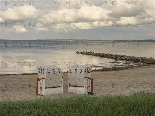 Two white beach chairs on a sandy beach, rocks in the water and cloudy sky in the background, beach