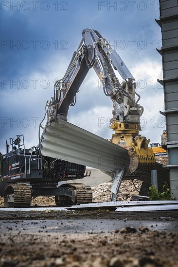 A large excavator lifts building materials on a construction site, the sky is cloudy and the