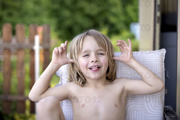 Boy, 5 years, blonde, sitting in garden chair, garden, grimacing, sticking out tongue, summer,