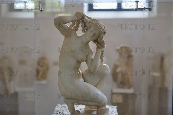 Aphrodite, crouching, Kneeling marble sculpture of a woman braiding her hair, surrounded by other