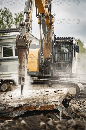 An excavator breaks up concrete on a construction site, steam rises, rubble and grey sky can be