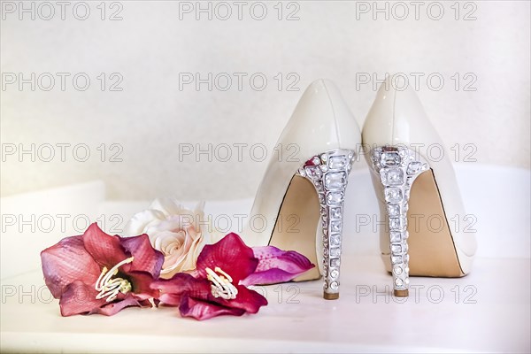 Fancy white high heels with rhinestone details next to pink flowers on a white bench creating an