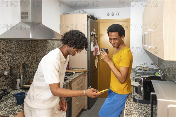 Gay couple laughing while using phone and drinking coffee in the morning in the kitchen at home
