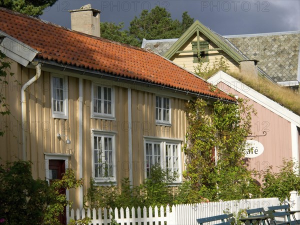 Yellow and pink house with a cafe sign and lush vegetation, colourful wooden houses behind white