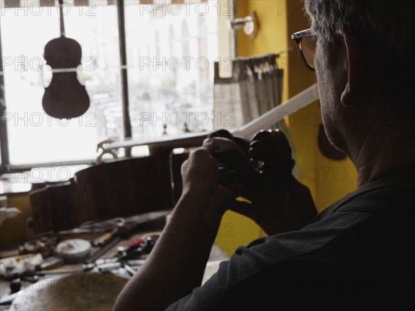 A luthier seen from behind works on a violin near a well-lit window, tools spread out on the bench