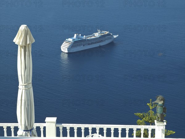 A cruise ship in the blue sea near a coastline with a parasol and railing in the foreground, The