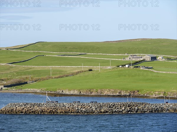 Small harbour landscape with green hills, houses and a stone embankment along the coast under a