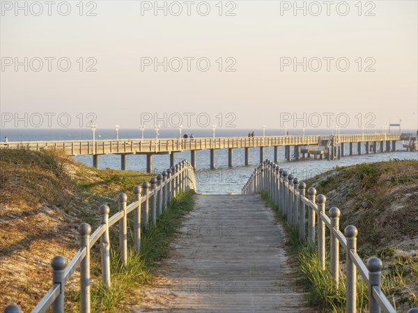 A long wooden walkway leads into the sea, framed by grass and promenade lighting in the soft