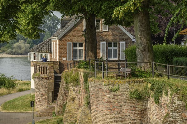 An old house on a riverbank, surrounded by a wall and trees, summery historical landscape, the