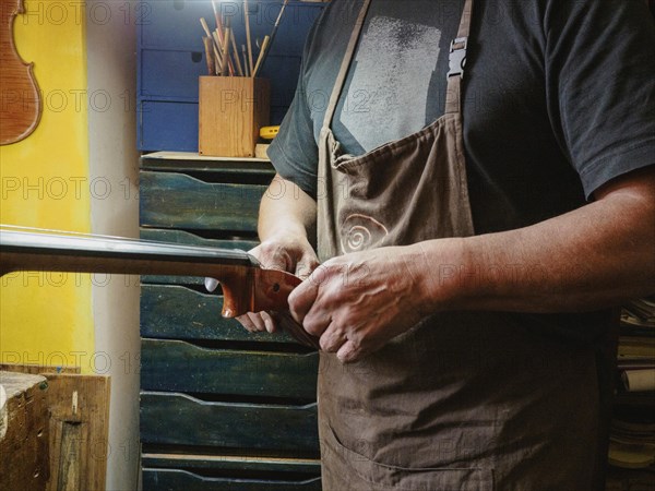 A luthier carefully works on a violoncello head and curl while wearing an apron, surrounded by