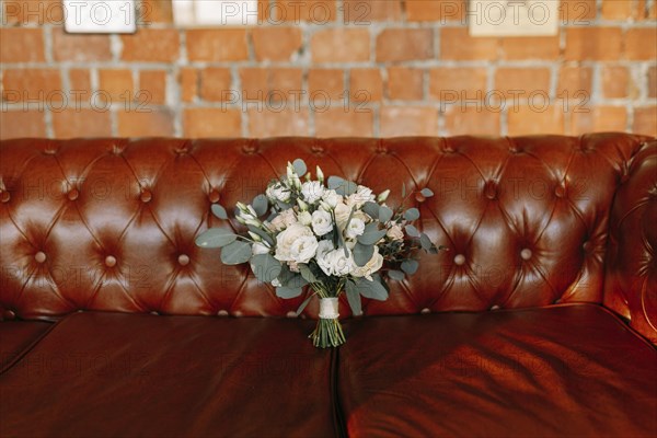 A bouquet of white flowers is placed on a brown leather couch against a brick wall
