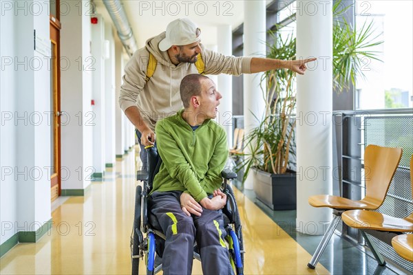 Caucasian young man showing something outside the university to a disabled friend sitting on