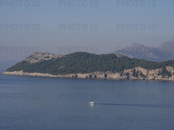 A small boat sails on the calm sea along a forested chain of islands, the old town of Dubrovnik