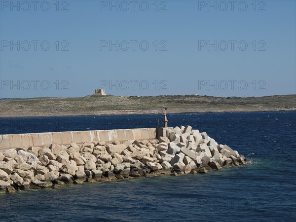 A breakwater of large stones in the sea with a rocky coastline in the background, the island of