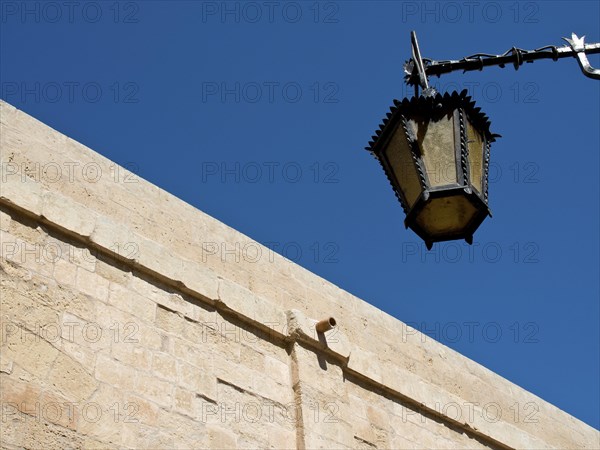 An ancient lantern hangs on a stone wall against a deep blue sky, the town of mdina on the island