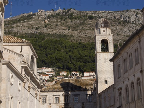 Church bell tower and historic buildings in front of a wooded mountain and blue sky, typical for
