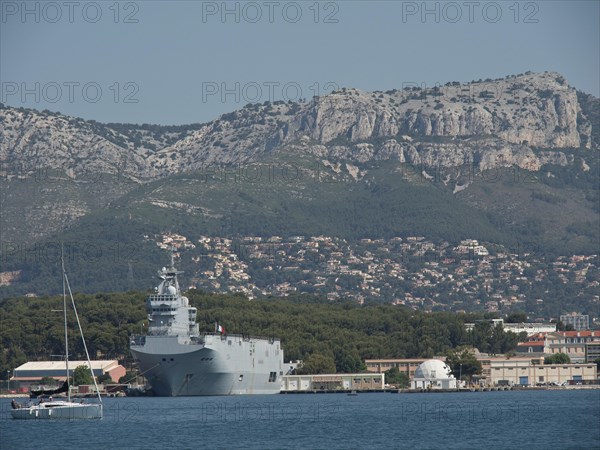 A warship lies next to a sailing boat in the harbour, with a mountain range in the background and a