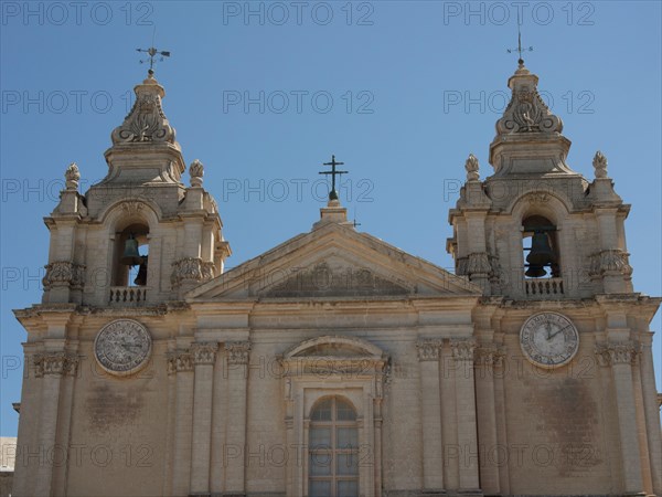 Front of a church with two heavily decorated bell towers and clocks, the town of mdina on the