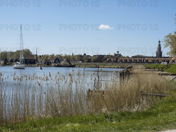 A lake with boats on the shore, a small town and a tower, reeds growing by the water, the scene